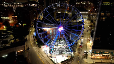 Aerial-flyover-illuminated-SkyView-Atlanta-Ferris-wheel-at-night-and-driving-Cars-on-road-in-downtown,-Georgia