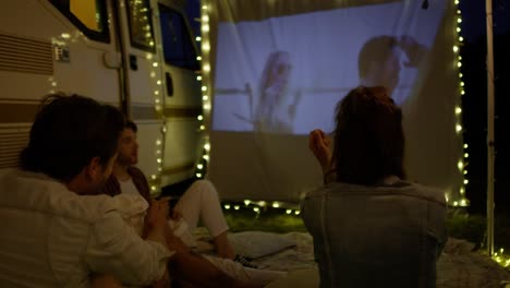 Group-of-young-cheerful-people-watching-a-movie-on-camping-site-on-projection-screen.