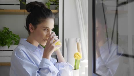 Young-woman-looking-through-window-and-drinking-orange-juice-at-home