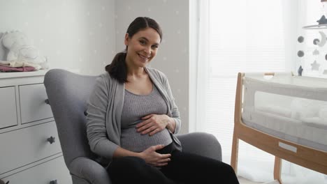 Portrait-of-woman-in-advanced-pregnancy-sitting-in-the-armchair-and-stroking-her-abdomen.