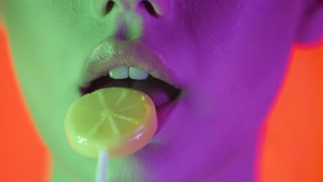 Extreme-Nahaufnahme-Der-Lippen-Einer-Jungen-Sexy-Frau,-Während-Sie-In-Zeitlupe-Einen-Süßen,-Köstlichen-Zitronenförmigen-Lutscher-Vor-Orangefarbenem-Hintergrund-Mit-Grün-violettem-Kontrast-Im-Gesicht-Dreht