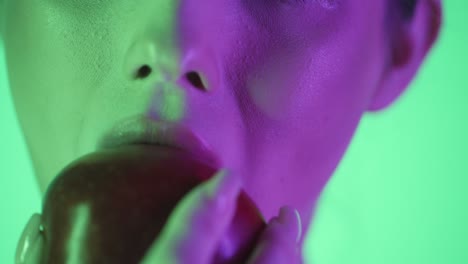 Extreme-Nahaufnahme-Der-Lippe-Einer-Frau,-Während-Sie-Vor-Grünem-Hintergrund-In-Zeitlupe-Einen-Angenehmen-Bissen-In-Einen-Roten-Apfel-Nimmt,-Mit-Grünem-Und-Violettem-Kontrast-In-Ihrem-Schönen-Gesicht