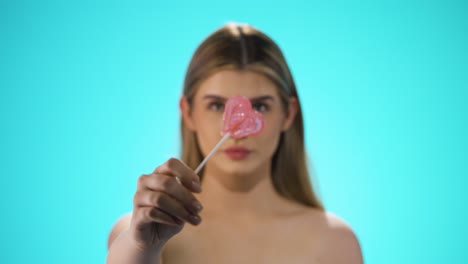 Medium-shot-of-a-young-beautiful-woman-holding-a-heart-shaped-delicious-lollipop-into-the-camera-in-front-of-turquoise-background-in-slow-motion