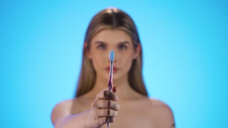 Static-medium-shot-of-a-young-beautiful-woman-or-model-holding-a-toothbrush-for-good-and-important-dental-care-and-dental-health-into-the-camera-in-front-of-blue-background-in-slow-motion