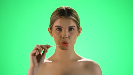 Medium-static-shot-of-a-young-pretty-woman-holding-a-heart-shaped-delicious-lollipop-into-the-camera-before-licking-it-and-enjoying-the-sweetness-in-front-of-green-background-in-slow-motion