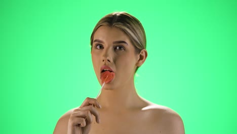 Medium-static-shot-of-a-young-pretty-woman-holding-a-heart-shaped-tasty-lollipop-into-the-camera-before-licking-it-and-enjoying-the-sweetness-in-front-of-green-background-in-slow-motion