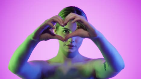 Medium-static-shot-of-a-pretty-young-woman-folding-her-hands-to-a-heart-symbolizing-love-and-mindfulness-in-front-of-pink-background-in-slow-motion