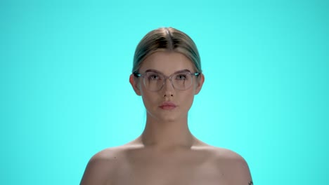 Mittlere-Statische-Aufnahme-Einer-Jungen,-Hübschen-Frau,-Die-Für-Eine-Klare-Sicht-Ihre-Brille-Aufsetzt-Und-In-Zeitlupe-Vor-Türkisfarbenem-Hintergrund-In-Die-Kamera-Blickt