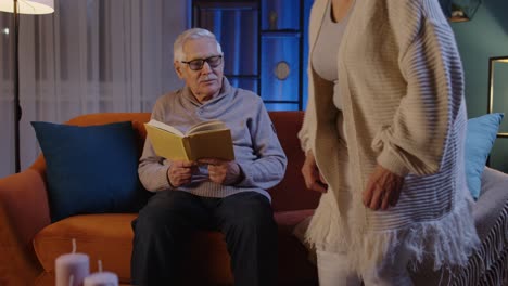 Senior-grandparents-couple-relaxing,-reading-book,-talking-enjoying-leisure-hobbies-at-evening-home