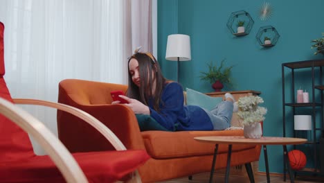 Relaxed-smiling-Caucasian-young-woman-lying-on-home-sofa-with-smartphone-scrolling,-browsing-online