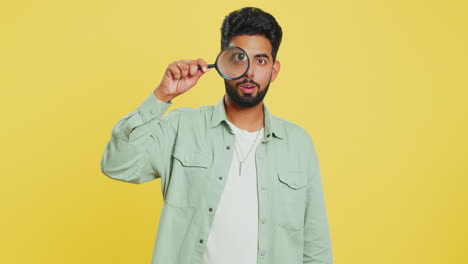Indian-young-man-holds-magnifying-glass-near-face-looking-at-camera-with-big-zoomed-eye,-analyzing