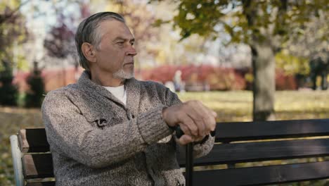Serious-old-caucasian-man-sitting-on-bench-in-park-during-the-autumn