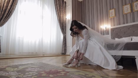 Young-bride-in-white-wedding-dress-wearing-high-heeled-shoes-on-slim-legs-while-sitting-at-home