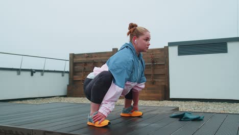 Sporty-fit-woman-in-blue-pink-sportswear-doing-squats-yoga-stretching-exercising-on-roof-of-house