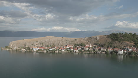 Droneshot-from-above-Lin-Albania-flying-backwards-on-a-cloudy-day-with-the-sun-coming-through-the-clouds-with-land-and-water-underneath-LOG