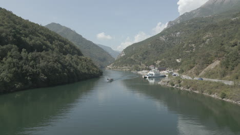 Disparo-De-Un-Dron-Sobrevolando-La-Parada-Del-Ferry-En-Fierze-Albania-Con-Un-Barco-Pasando-Y-Una-Parada-Del-Ferry-En-El-Agua-En-Un-Día-Soleado-Y-Nublado