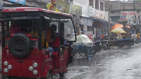 Rural-Indian-town-during-rainfall,-Locals-traveling-in-electronic-rickshaw-and-tuk-tuk-with-the-view-of-household-goods-shops-in-the-background