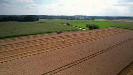 Aerial-backwards-shot-of-tractor-harvesting-corn-of-farm-field-in-countryside-at-summer-season---forest-woodland-in-backdrop