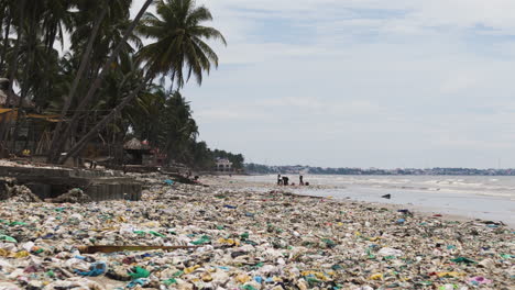 Tropical-Beach-Polluted-with-Plastic-Trash,-Locals-Cleaning-Up-in-the-Distance
