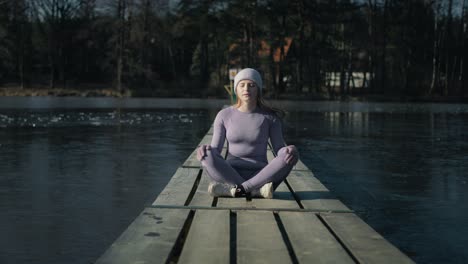 Zoom-out-of-caucasian-woman-sitting-on-pier-and-meditating-with-eyes-closed-in-the-winter.