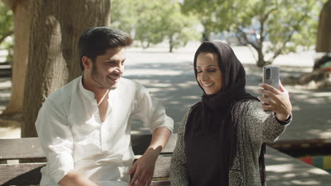 Happy-muslim-couple-sitting-on-bench,-making-selfies.