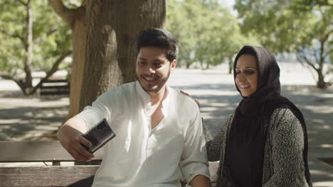 Young-muslim-couple-sitting-on-bench-in-park,-taking-selfie.
