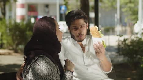 Junges-Muslimisches-Paar-Sitzt-Auf-Einer-Bank-Und-Isst-An-Einem-Sonnigen-Tag-Einen-Snack.
