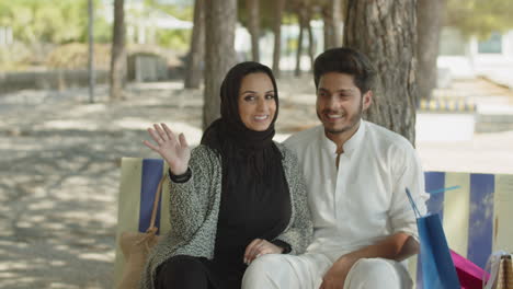 Young-muslim-couple-sitting-on-bench-in-park,-waving-to-camera.