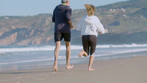 Elderly-couple-running-barefoot-on-sandy-beach-and-having-fun