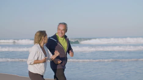 Cheerful-mature-couple-running-along-ocean-coast-in-morning