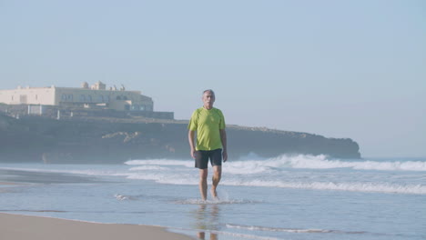 Tired-senior-man-in-sportswear-walking-barefoot-on-sandy-beach