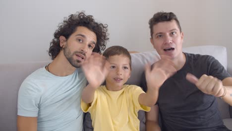 Two-happy-dads-and-child-waving-hello-at-camera