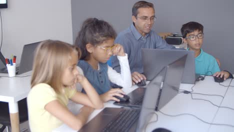 Computer-science-teacher-sitting-at-desk-near-children