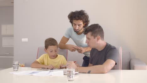 Dos-Papás-Ayudando-A-Un-Niño-Concentrado-Con-La-Tarea-De-La-Escuela-En-Casa