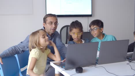 Computer-science-teacher-sitting-at-desk-near-children