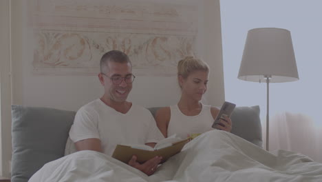 Man-reading-book-in-bed,-woman-looking-at-phone-screen-at-home
