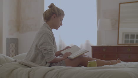 Focused-woman-sitting-on-bed-and-reading-book-after-waking-up