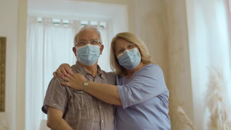 Senior-couple-in-medical-mask-standing-at-home-and-hugging