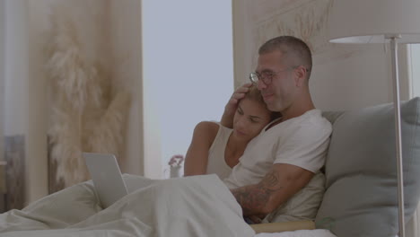 Happy-man-and-woman-sitting-on-bed-and-watching-movie-on-laptop