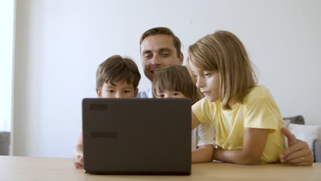 Loving-dad-sitting-at-table,-embracing-kids-and-using-laptop