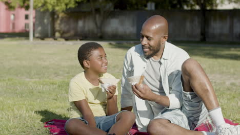 Black-father-and-son-having-picnic-in-park,-eating-sandwiches.