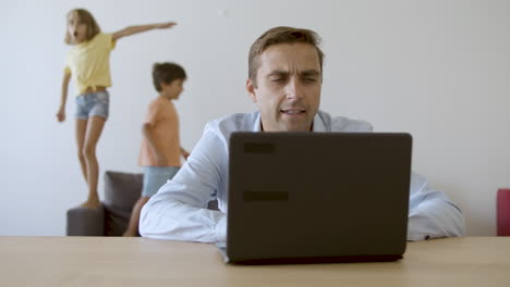 Serious-dad-working-via-laptop-and-kids-jumping-on-background