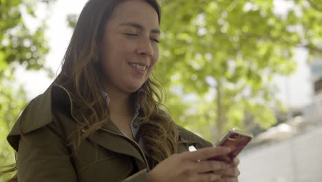Mujer-Joven-Sonriente-Sosteniendo-Un-Moderno-Teléfono-Inteligente-Al-Aire-Libre
