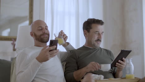 Happy-gay-couple-having-breakfast-in-bed-and-using-gadgets