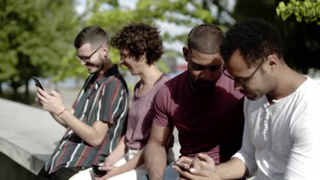 Happy-friends-sitting-in-park-and-using-smartphones