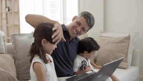 Cheerful-dad-and-two-cute-kids-sitting-at-laptop-in-living-room