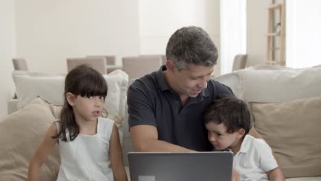 Cheerful-dad-and-two-cute-kids-sitting-on-couch-at-laptop