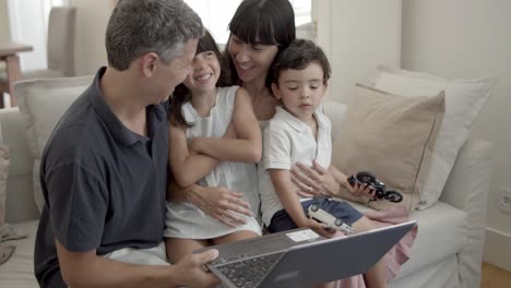 Joyful-parents-and-two-children-sitting-with-laptop-on-couch