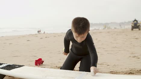Adorable-child-waxing-surfboard-on-beach