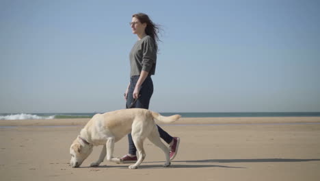 Relaxed-woman-in-eyeglasses-strolling-with-dog-on-seashore